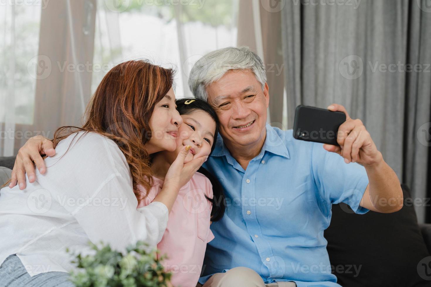 autofoto de abuelos asiáticos con nieta en casa. chino senior, abuelo y abuela felices pasan tiempo en familia relajarse usando el teléfono móvil con un niño niña acostado en el sofá en el concepto de sala de estar. foto