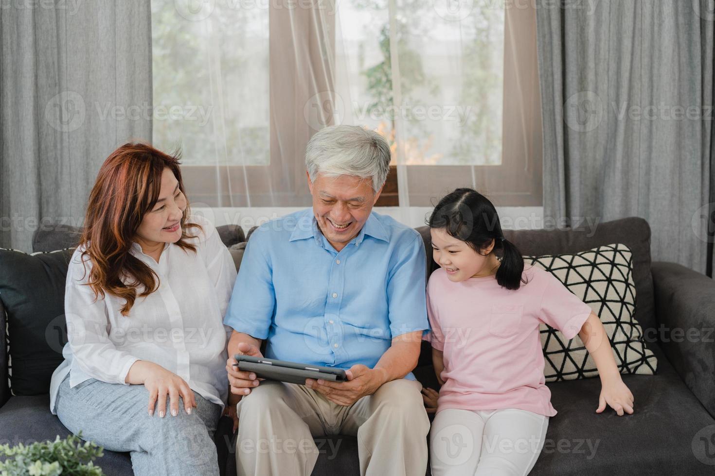 Asian grandparents and granddaughter using tablet at home. Senior Chinese, grandpa and grandma happy spend family time relax with young girl checking social media, lying on sofa in living room concept photo