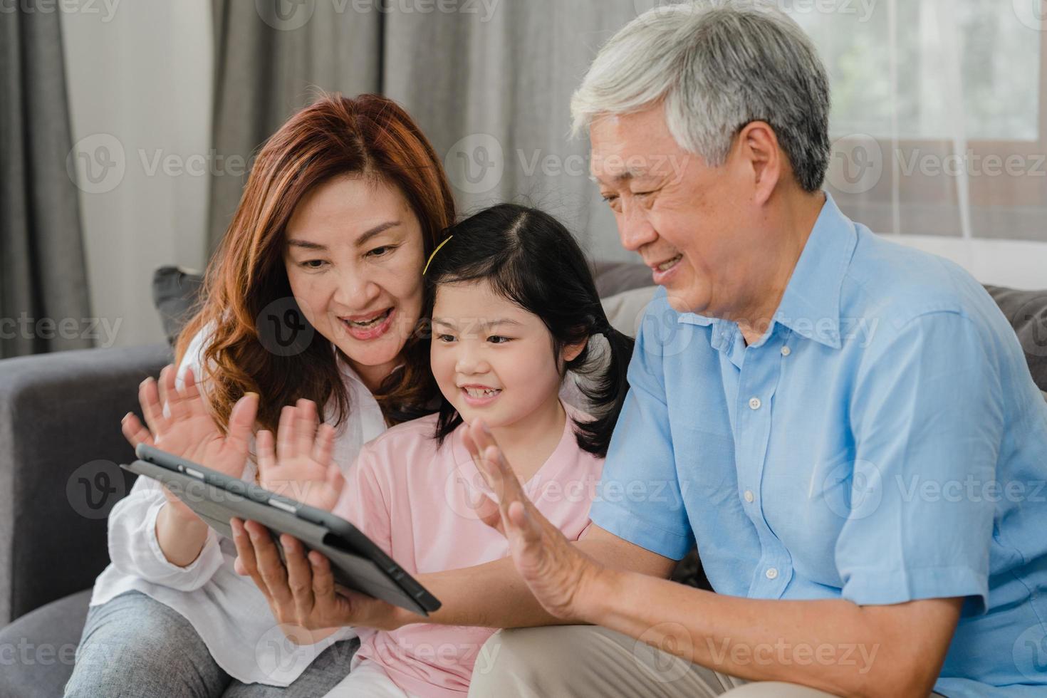 Asian grandparents and granddaughter video call at home. Senior Chinese, grandpa and grandma happy with girl using mobile phone video call talking with dad and mom lying in living room at home. photo