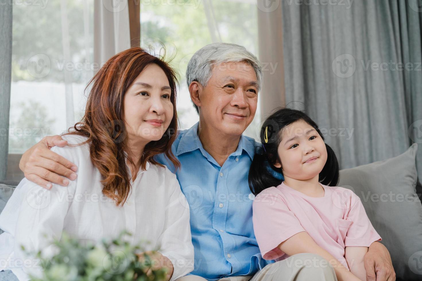 los abuelos asiáticos ven la televisión con su nieta en casa. chino senior, abuelo y abuela felices usando tiempo en familia relajarse con un niño niña acostado en el sofá en el concepto de sala de estar. foto