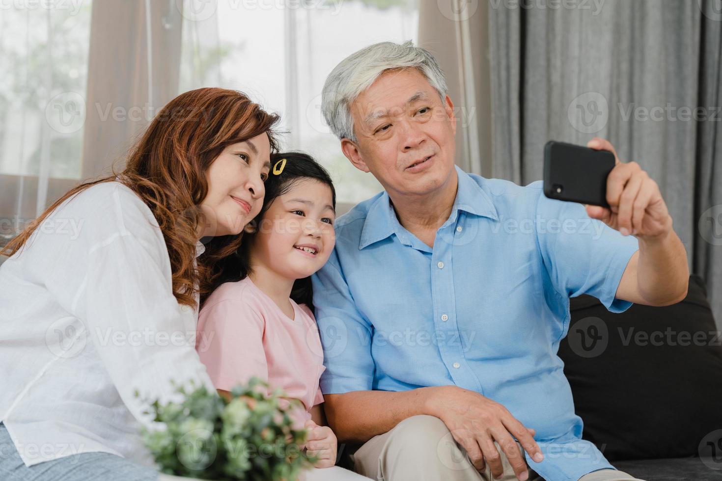 Asian grandparents selfie with granddaughter at home. Senior Chinese, grandpa and grandma happy spend family time relax using mobile phone with young girl kid lying on sofa in living room concept. photo