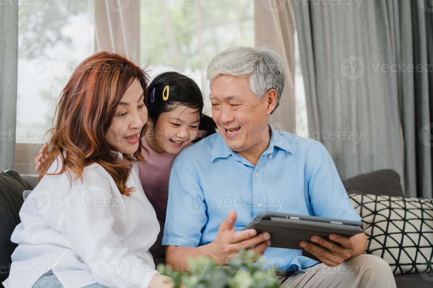 Asian grandparents and granddaughter using tablet at home. Senior Chinese, grandpa and grandma happy spend family time relax with young girl checking social media, lying on sofa in living room concept photo