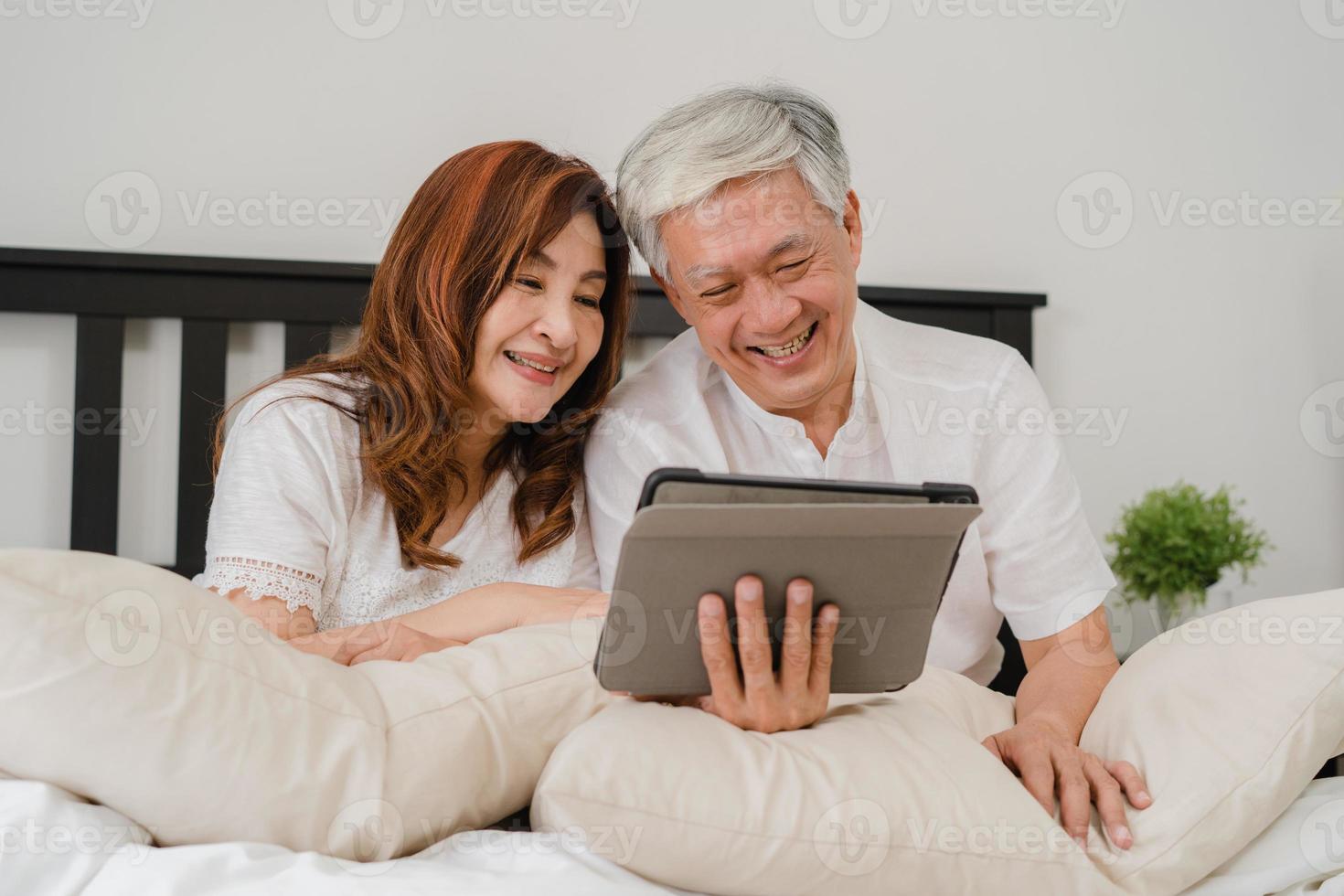 Asian senior couple using tablet at home. Asian Senior Chinese grandparents, husband and wife happy after wake up, watching movie lying on bed in bedroom at home in the morning concept. photo