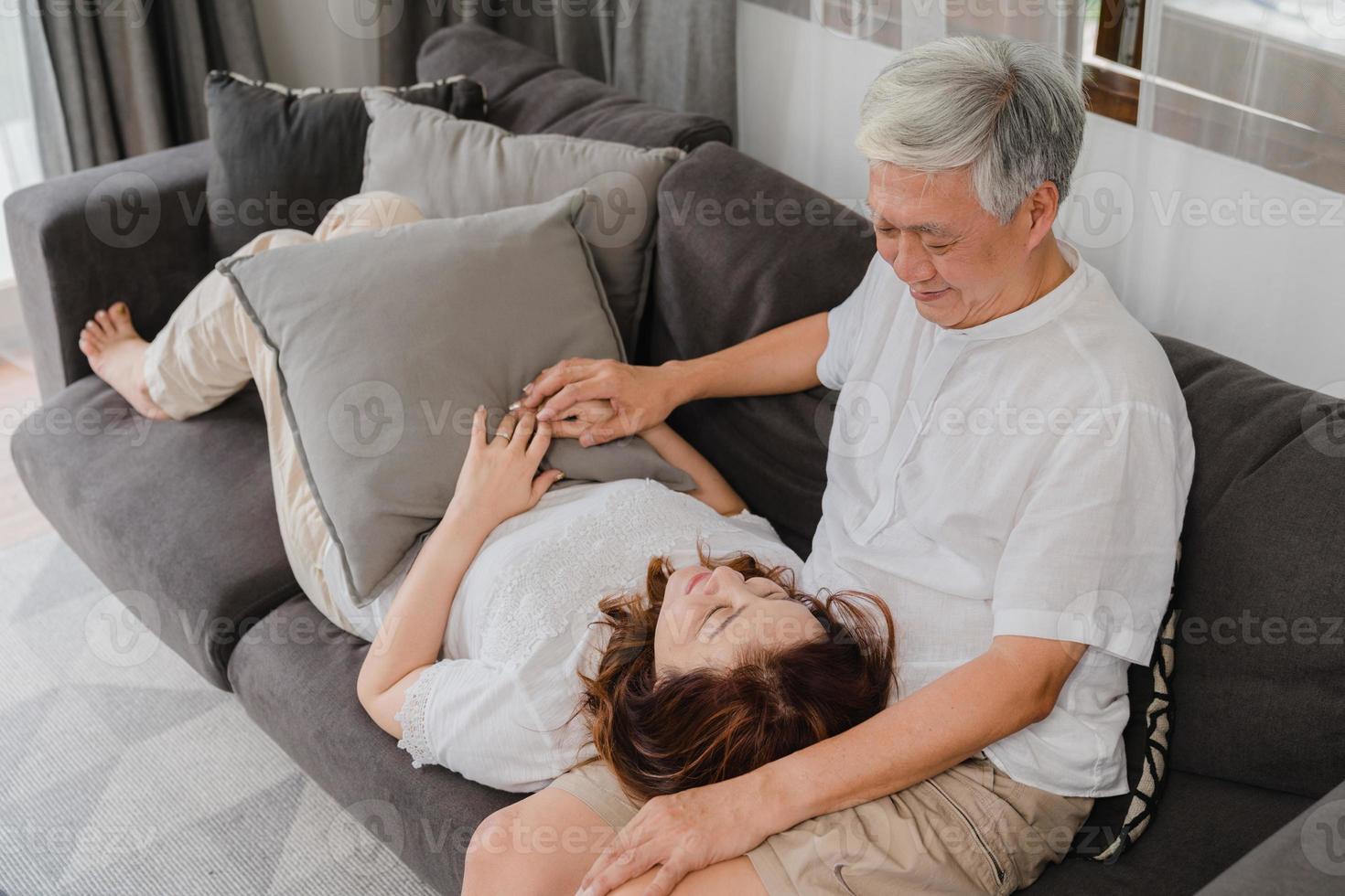 pareja senior asiática relajarse en casa. Abuelos chinos mayores asiáticos, abrazo feliz de la sonrisa del marido acostarse en el regazo de su esposa mientras está acostado en el sofá en la sala de estar en el concepto de casa. foto