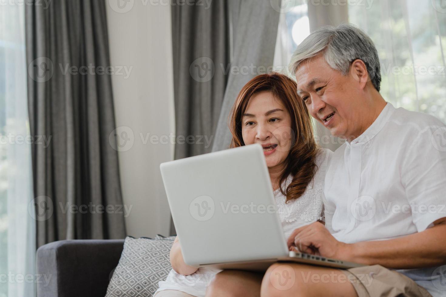 Asian senior couple using laptop at home. Asian Senior Chinese grandparents, surf the Internet to check social media while lying on sofa in living room at home concept. photo