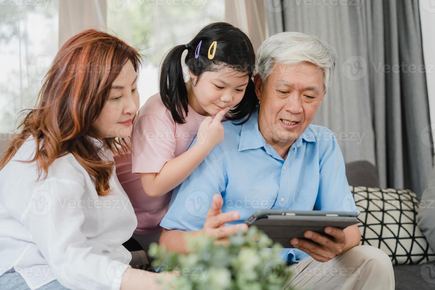 Asian grandparents and granddaughter using tablet at home. Senior Chinese, grandpa and grandma happy spend family time relax with young girl checking social media, lying on sofa in living room concept photo