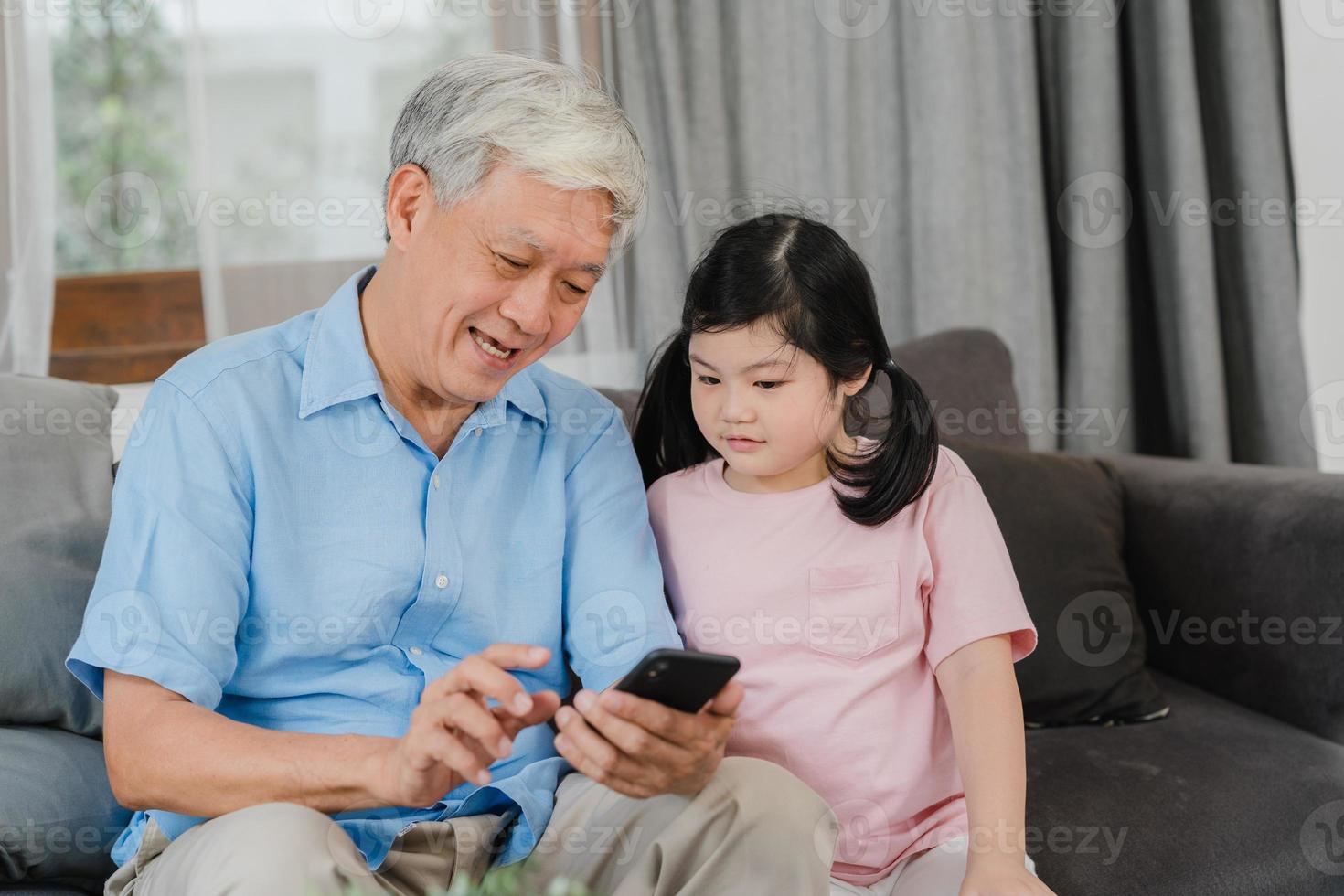 Asian grandparents and granddaughter using mobile phone at home. Senior Chinese, grandpa and kid happy spend family time relax with young girl checking social media, lying on sofa in living room. photo