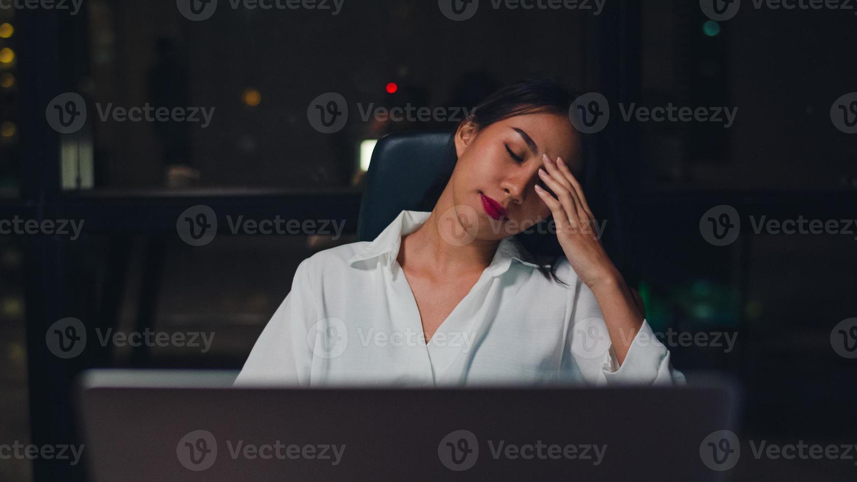 Joven empresaria china milenaria trabajando hasta altas horas de la noche con estrés con un problema de investigación del proyecto en una computadora portátil en la sala de reuniones de una pequeña oficina moderna. concepto de síndrome de agotamiento ocupacional de personas de Asia. foto