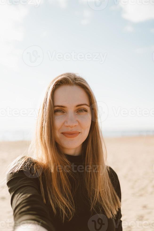 Portrait of pretty female with long hair, blonde takes selfie on mobile phone on sandy beach photo