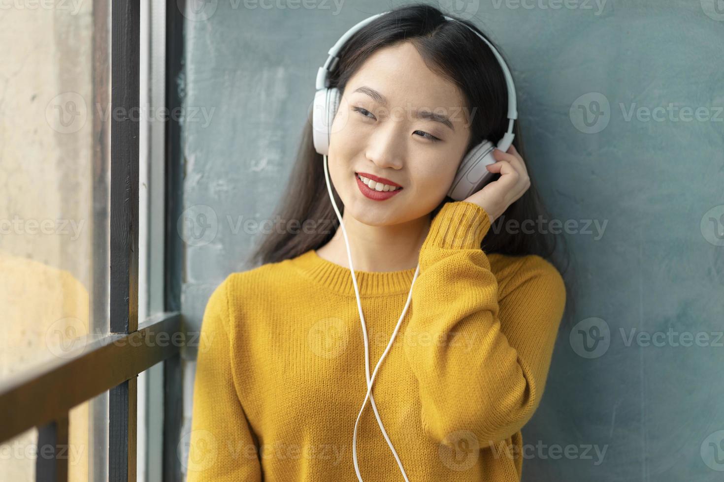 Hermosa joven asiática escuchando música con auriculares sentado en la ventana foto