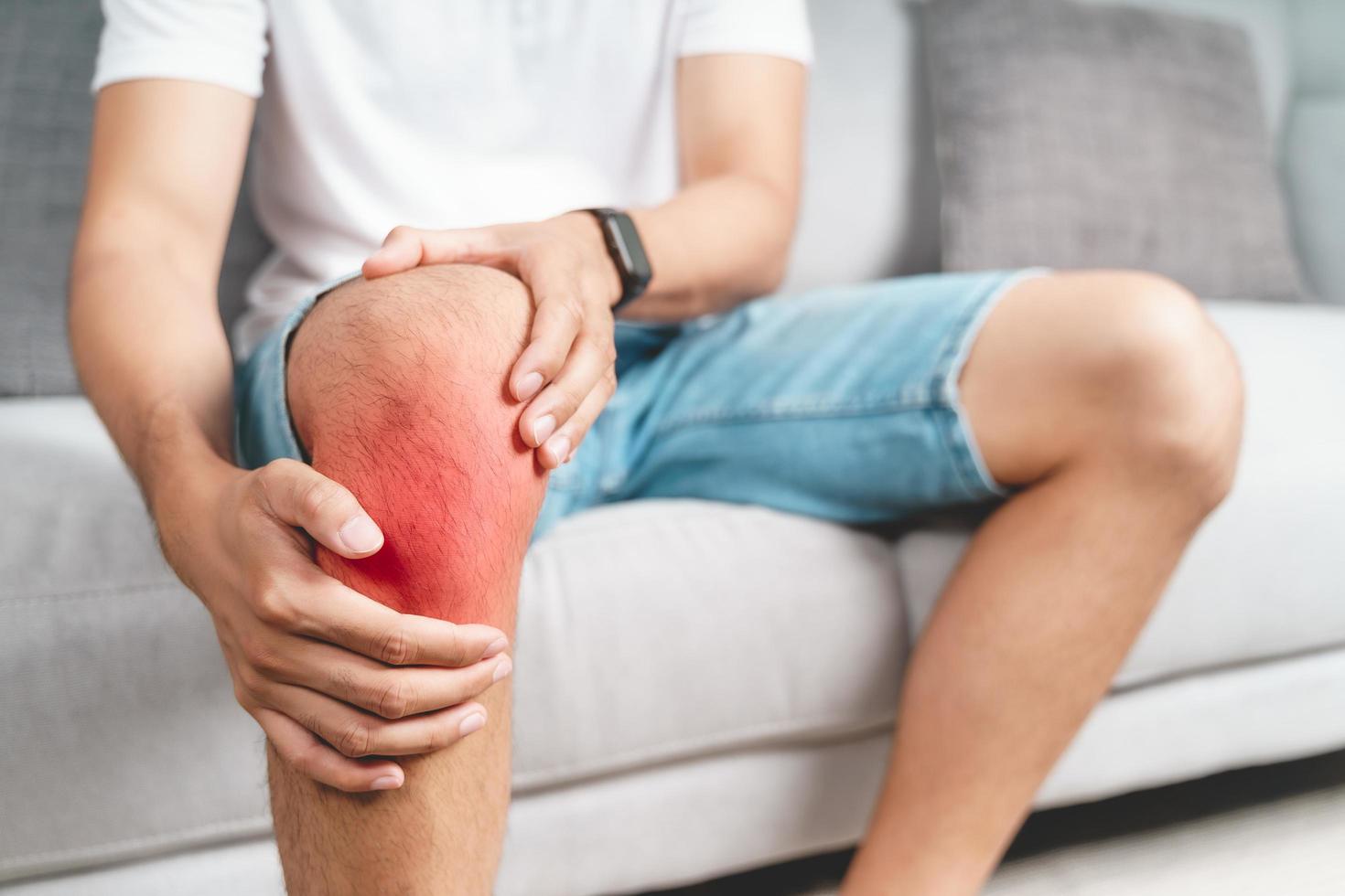 el hombre que sufre de dolor de rodilla sentado en el sofá sosteniendo y masajeando su rodilla dolorida. foto