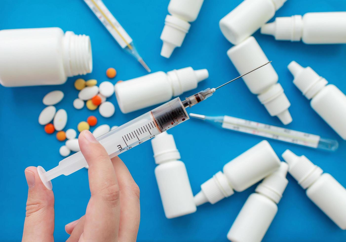 Mano sosteniendo una jeringa con medicamento en el fondo de gotas nasales, termómetros y píldoras sobre un fondo azul. concepto de protección de la salud. foto