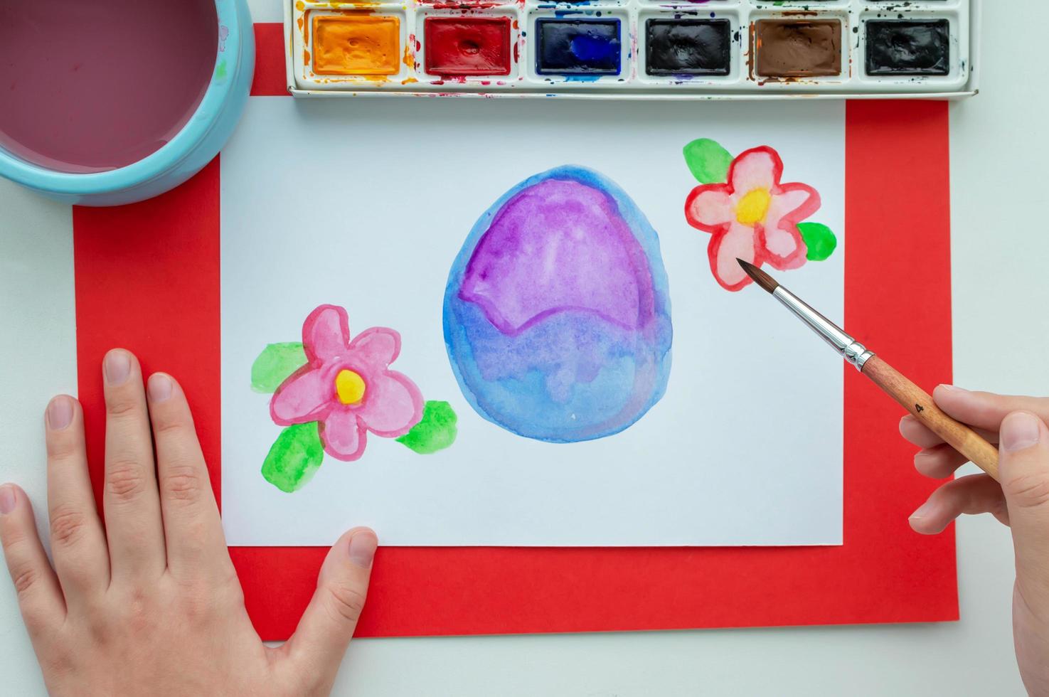 La mano del niño dibuja un huevo de pascua y flores en acuarela sobre una sábana blanca. foto