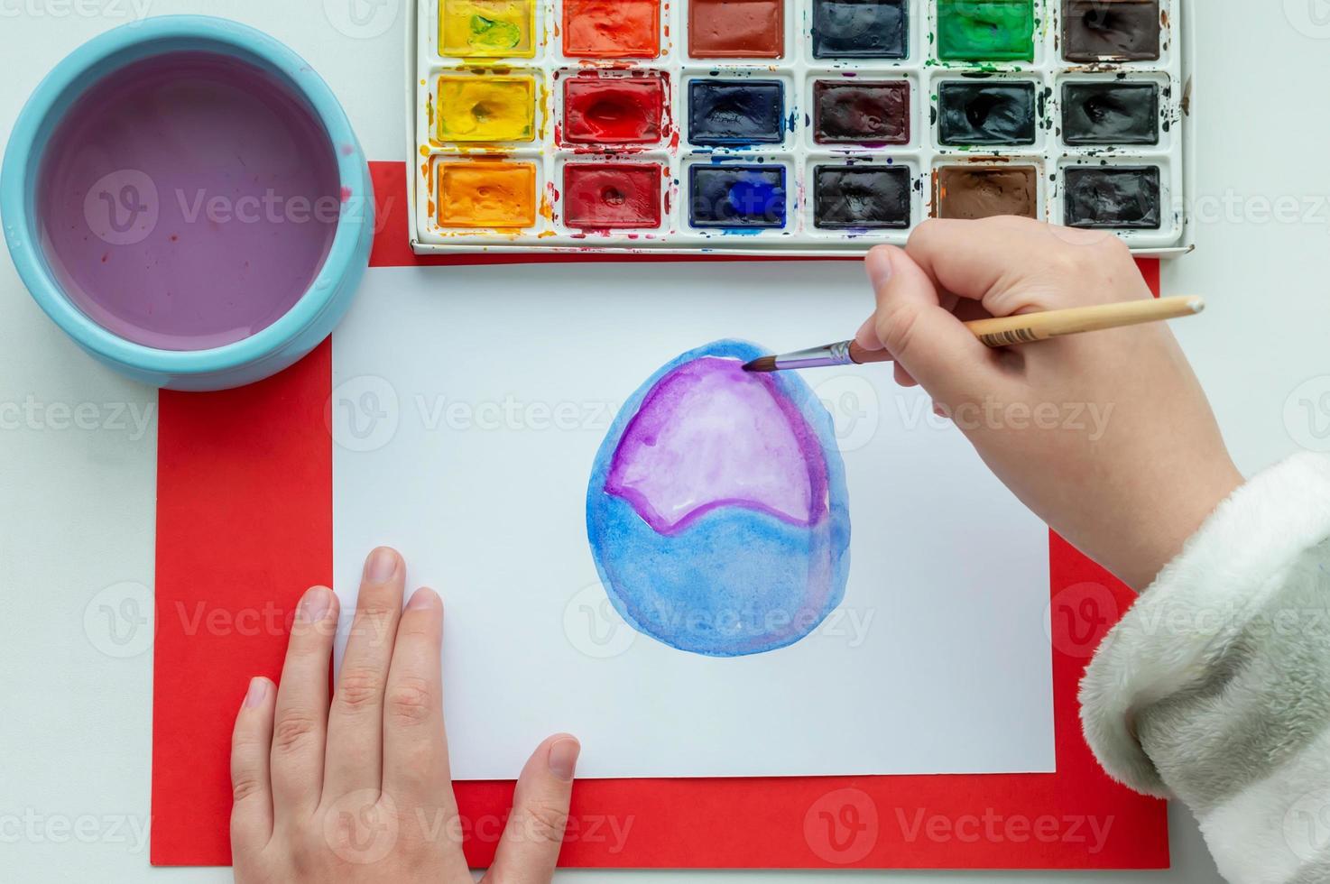 The child's hand draws an Easter egg and flowers in watercolor on a white sheet. photo