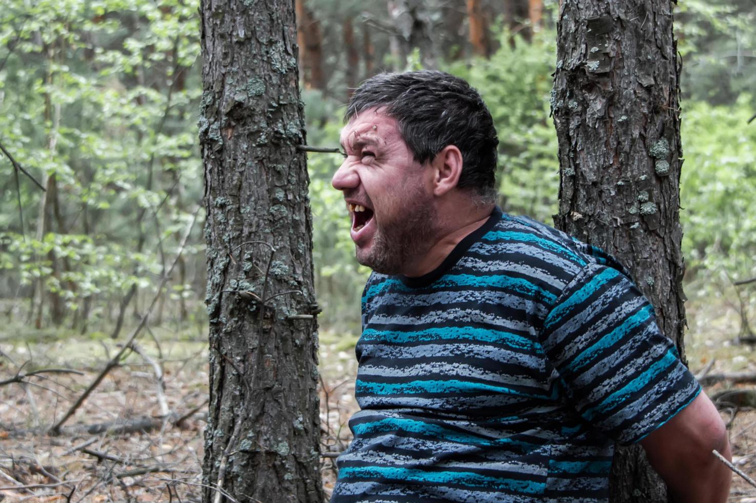 A hostage in the woods. A man in a blue t-shirt and trousers is sitting on the ground tied to a tree in the forest calling for help. Victim of an attack. Horizontal photo. photo