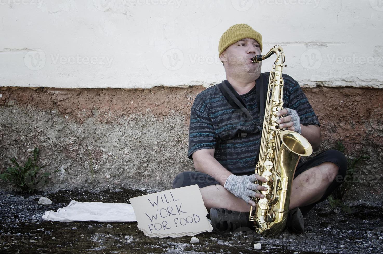 un músico callejero sin hogar se sienta en la calle con un saxofón para ganarse la comida. foto