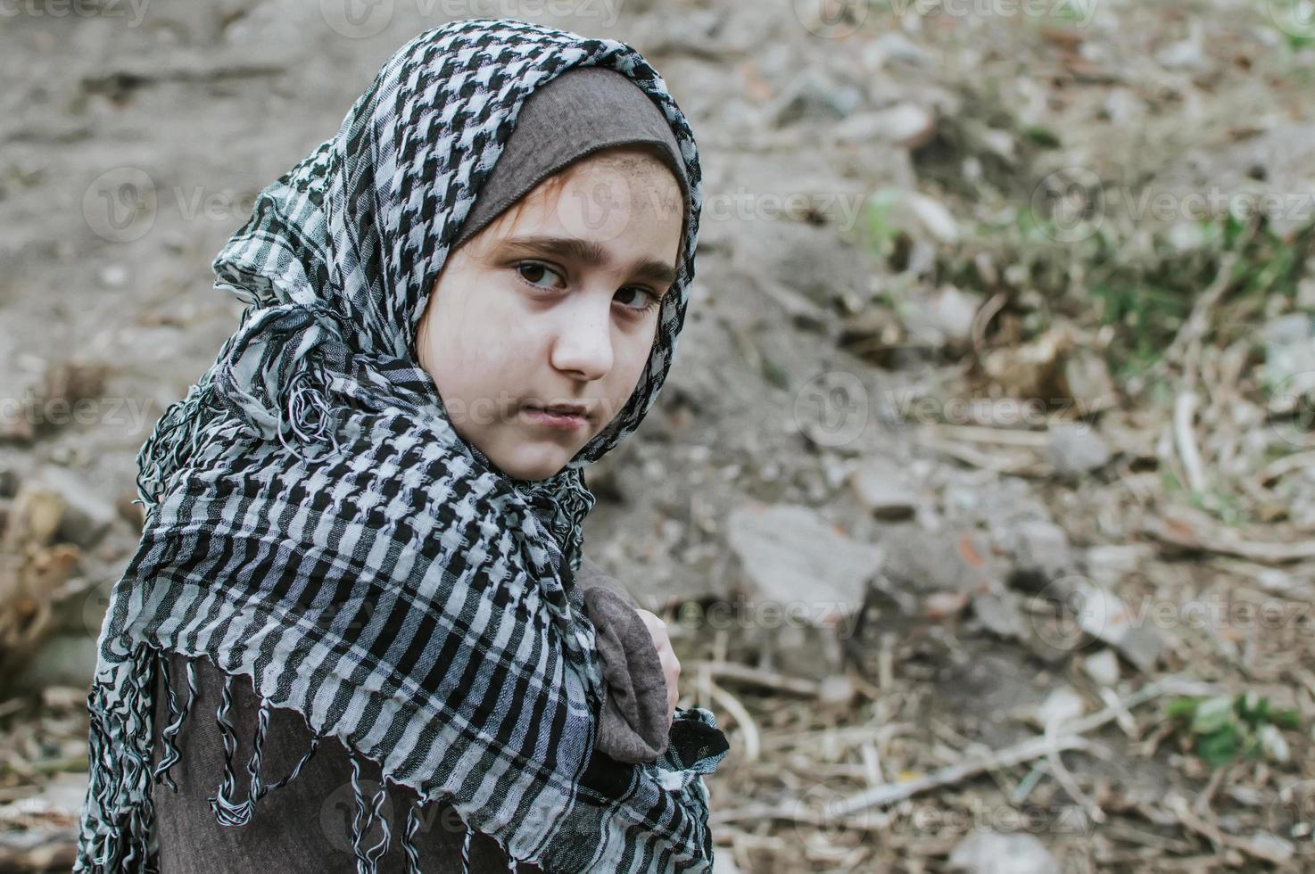a refugee child in the war, a Muslim girl with a dirty face on the ruins, the concept of peace and war, the child is crying and waiting for help. photo