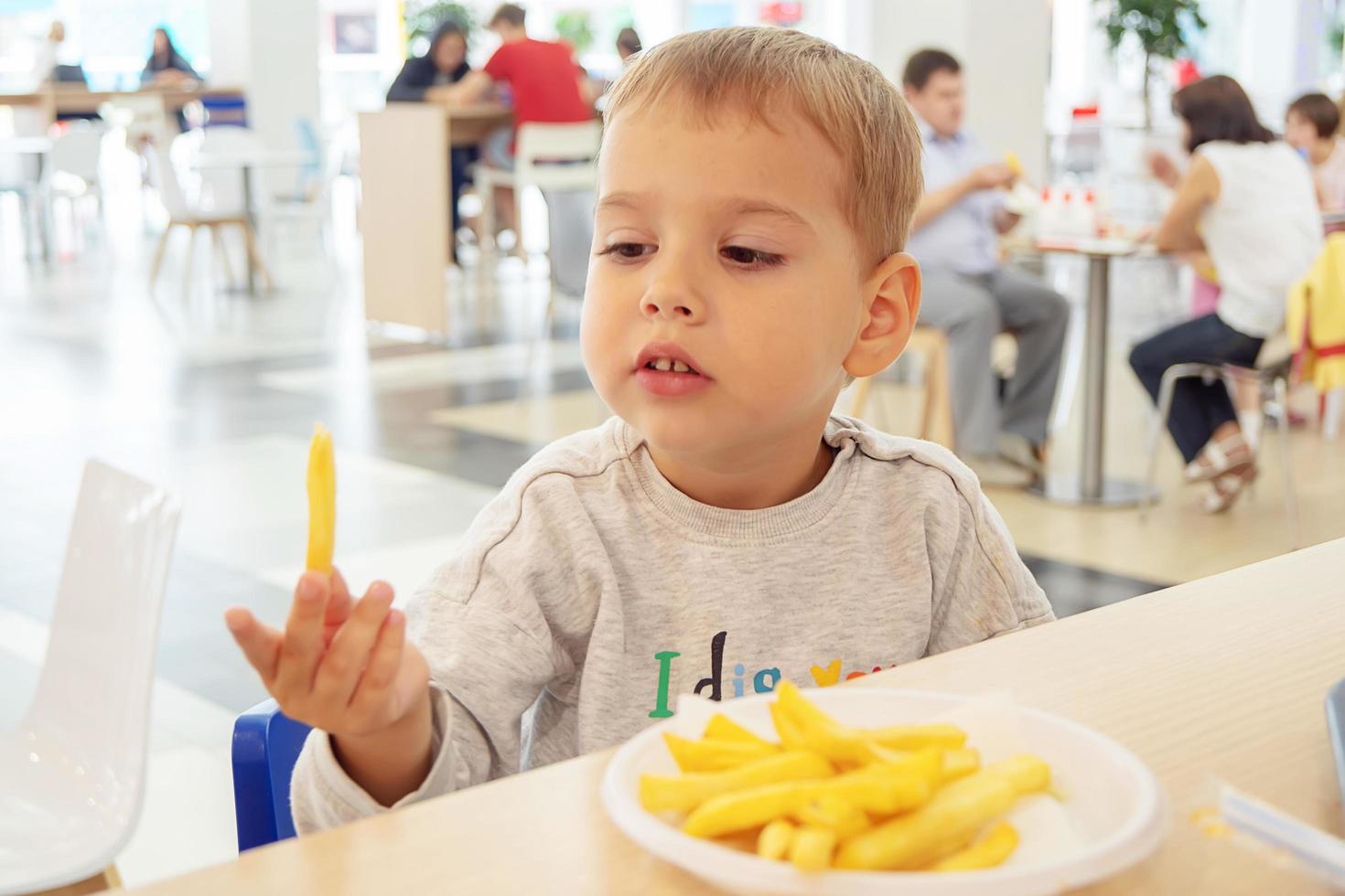 little child eating french fries sitting at a table on the food court of the mall. Unhealthy food. photo