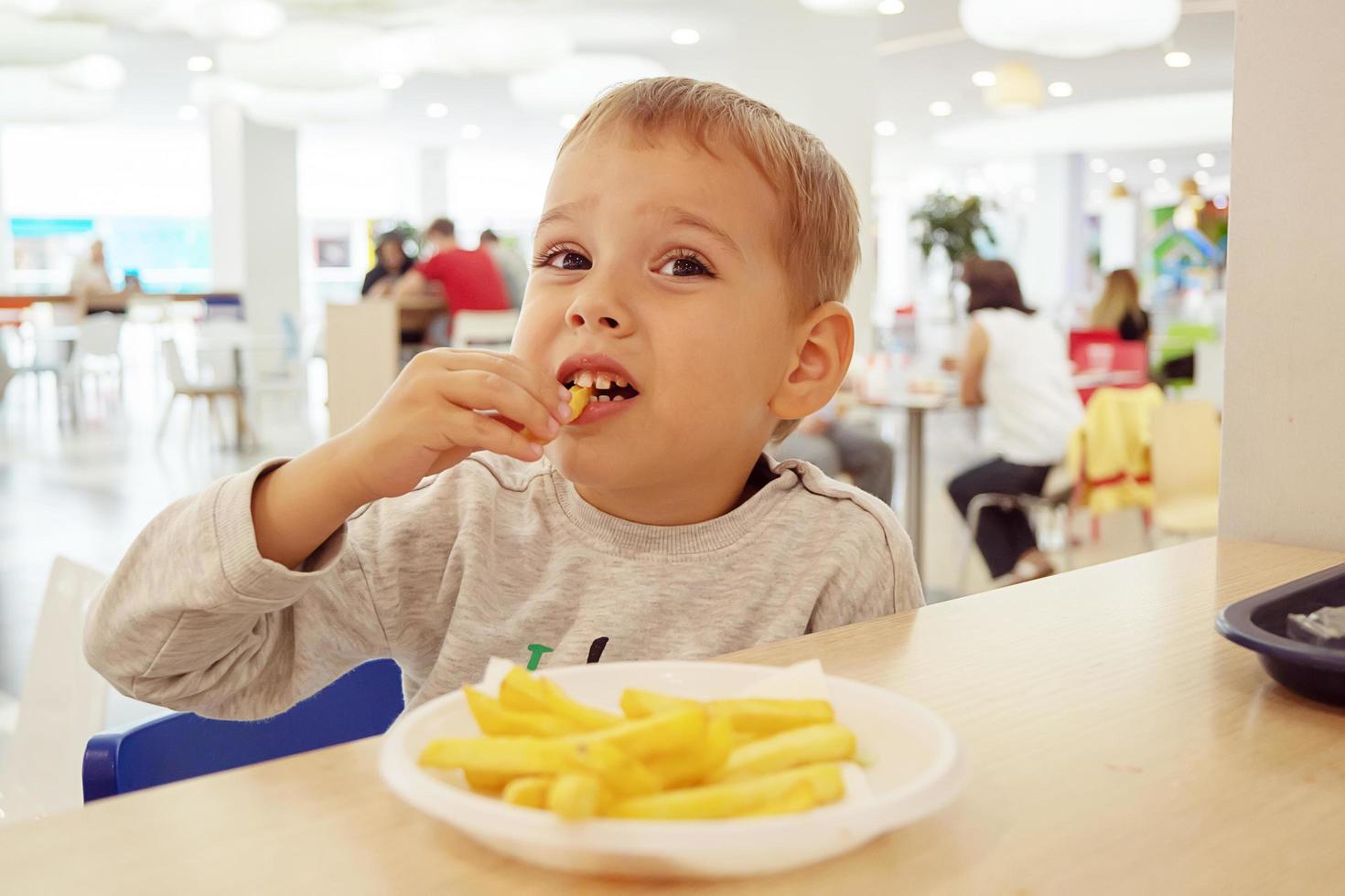 niño comiendo papas fritas sentado en una mesa en el patio de comidas del centro comercial. comida chatarra. foto