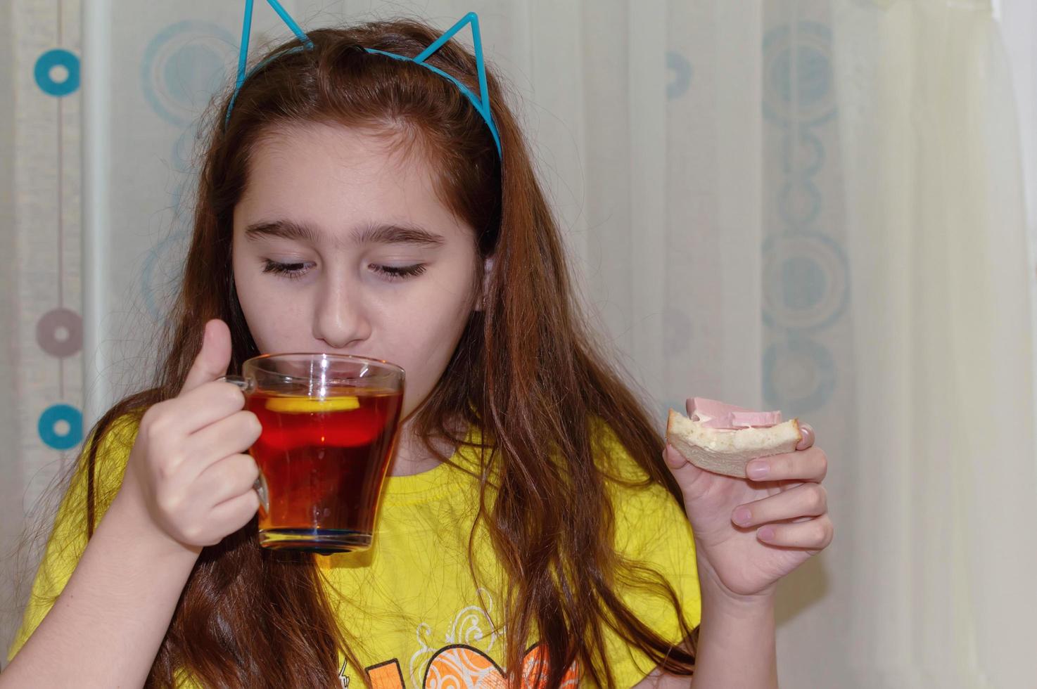 a teenage girl is sitting at a table in the kitchen and drinking tea with a sandwich. Morning, Breakfast, junk food. photo