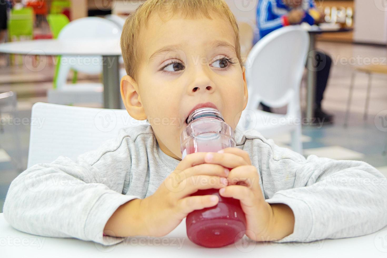 little child drinks juice sitting at a table on the food court of the mall. photo