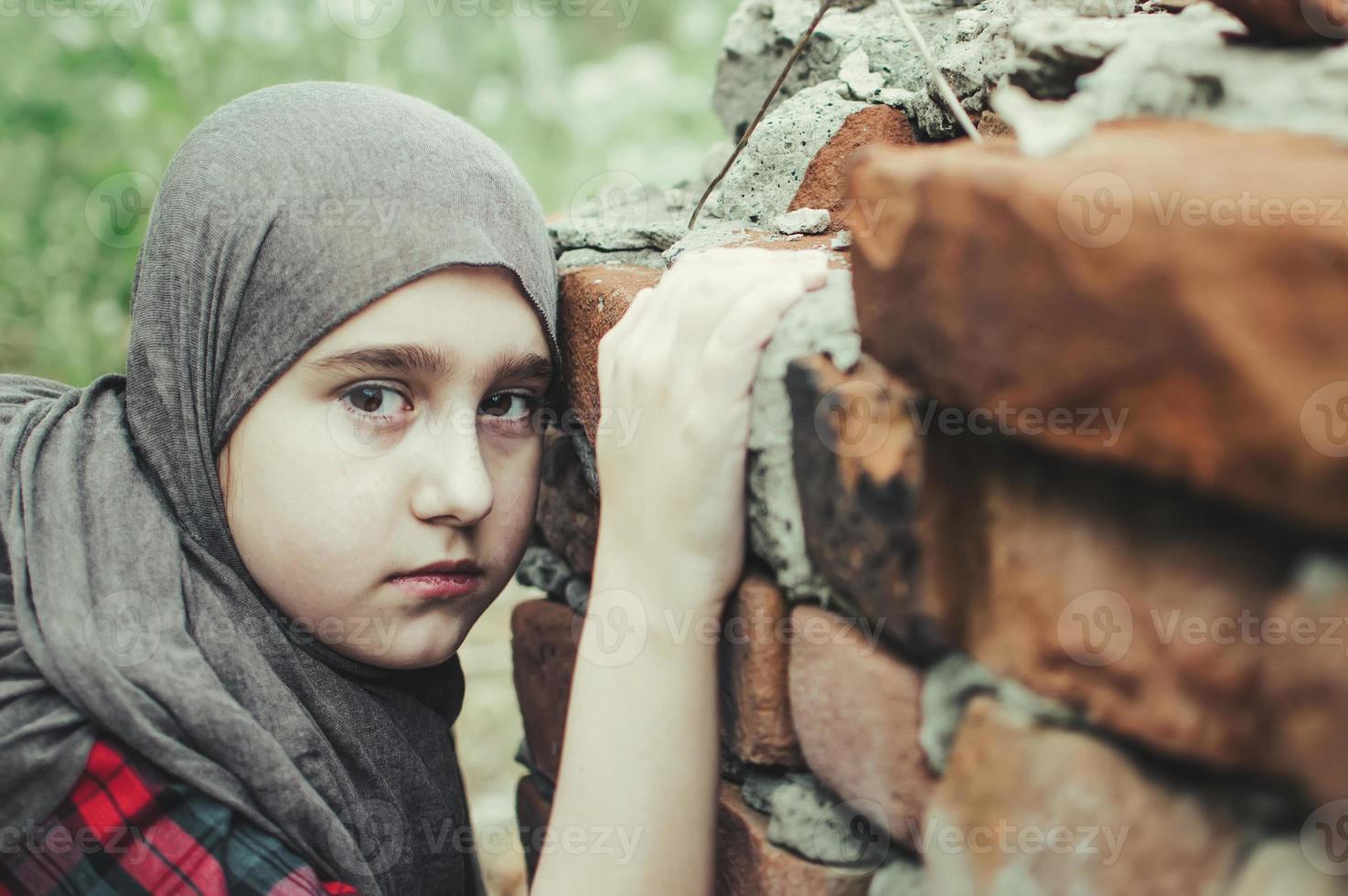 a refugee child in the war, a Muslim girl with a dirty face on the ruins, the concept of peace and war, the child is crying and waiting for help. photo