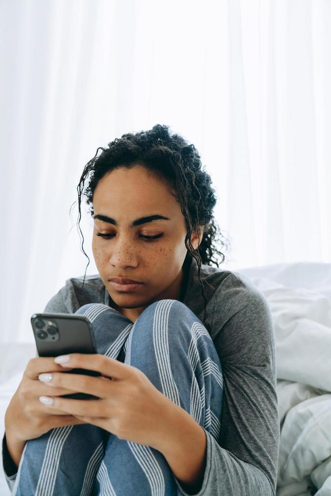 Mujer africana concentrada enviando mensajes de texto por teléfono en la cama foto