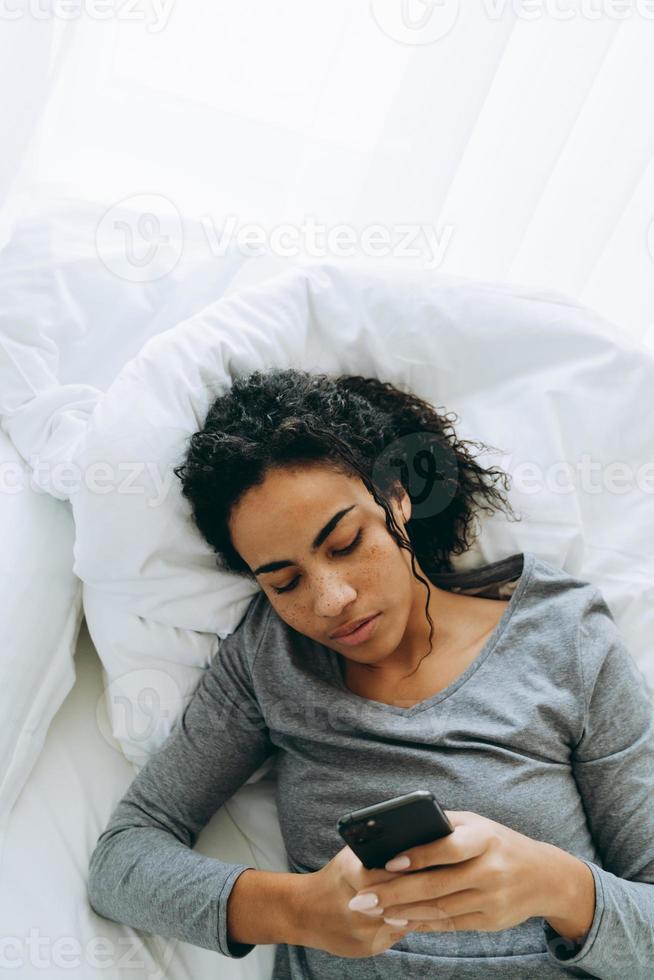 Young black woman using mobile phone while lying in her bed at home photo