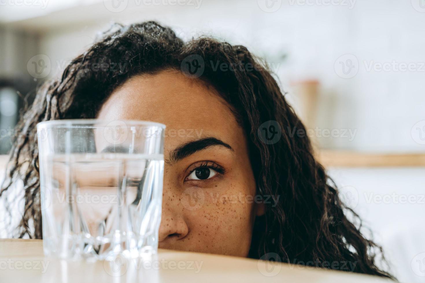 Joven mujer negra mirando el vaso de agua mientras está sentado en la cocina foto