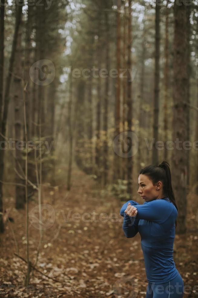 Mujer joven en traje de pista azul que se extiende antes de entrenar en el bosque de otoño foto