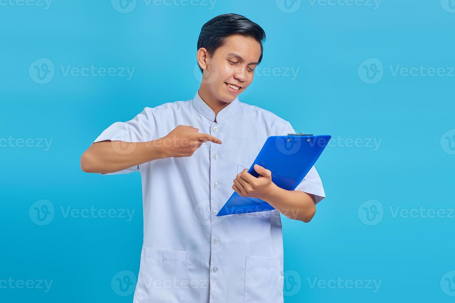 retrato, de, asiático, enfermero, señalar con el dedo, en, portapapeles, y, sonriente, en, fondo azul foto