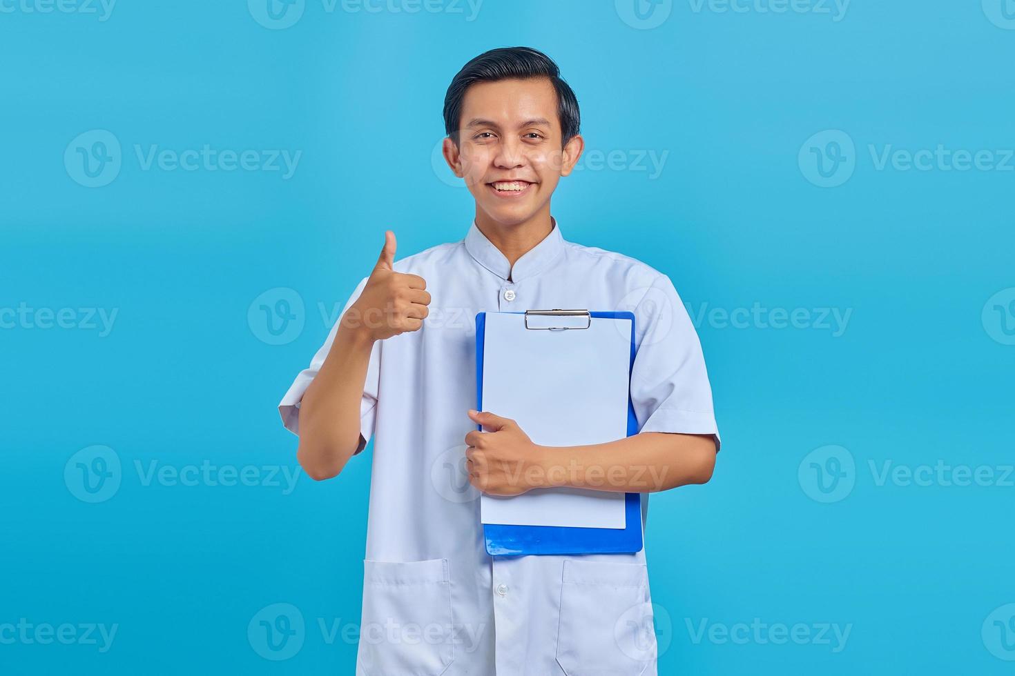 Sonriente joven enfermero mostrando el portapapeles y el pulgar hacia arriba gesto sobre fondo azul. foto