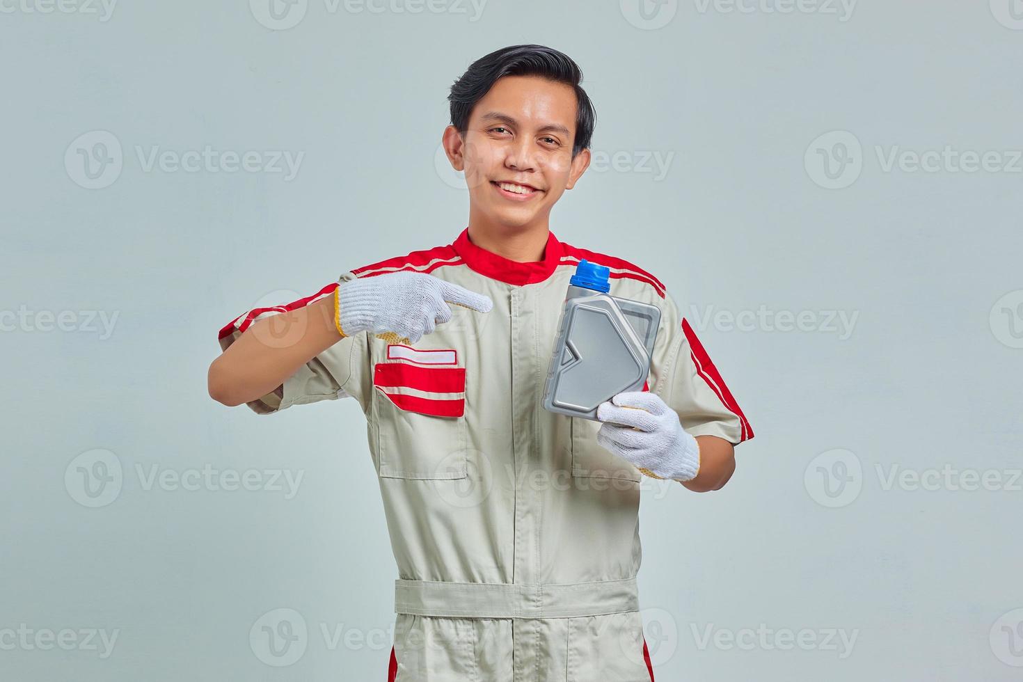 Retrato de alegre apuesto hombre vestido con uniforme de mecánico mostrando botella de plástico de aceite de motor sobre fondo gris foto