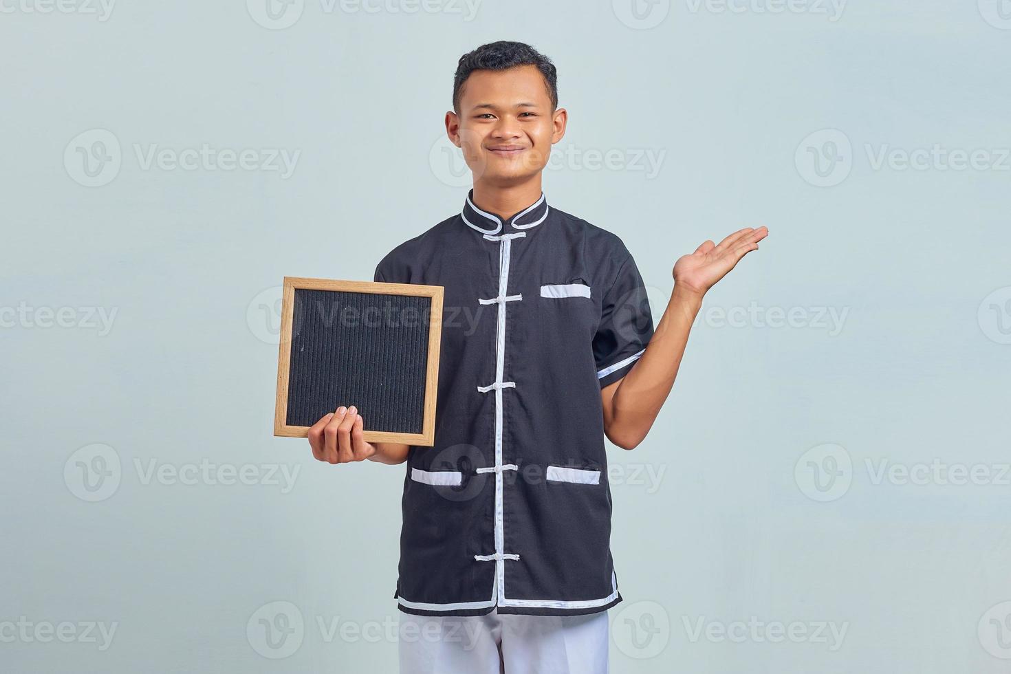 Portrait of cheerful Asian young man wearing karate uniform pointing to copy room with palms and holding blank board on gray background photo