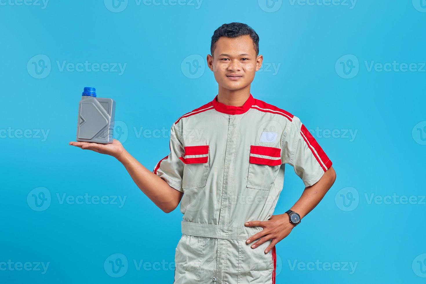 Retrato de smilling joven mecánico asiático mostrando botella de plástico de aceite de motor en la palma de la mano sobre fondo azul. foto