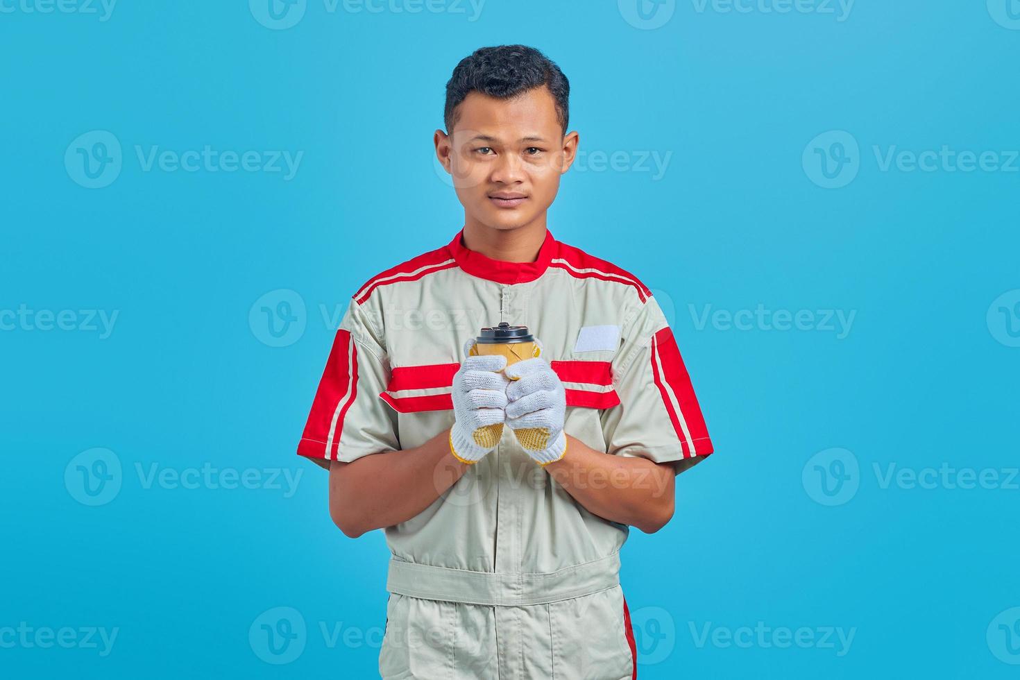 Retrato de sonriente joven mecánico asiático llevando una taza de café en la mano sobre fondo azul. foto