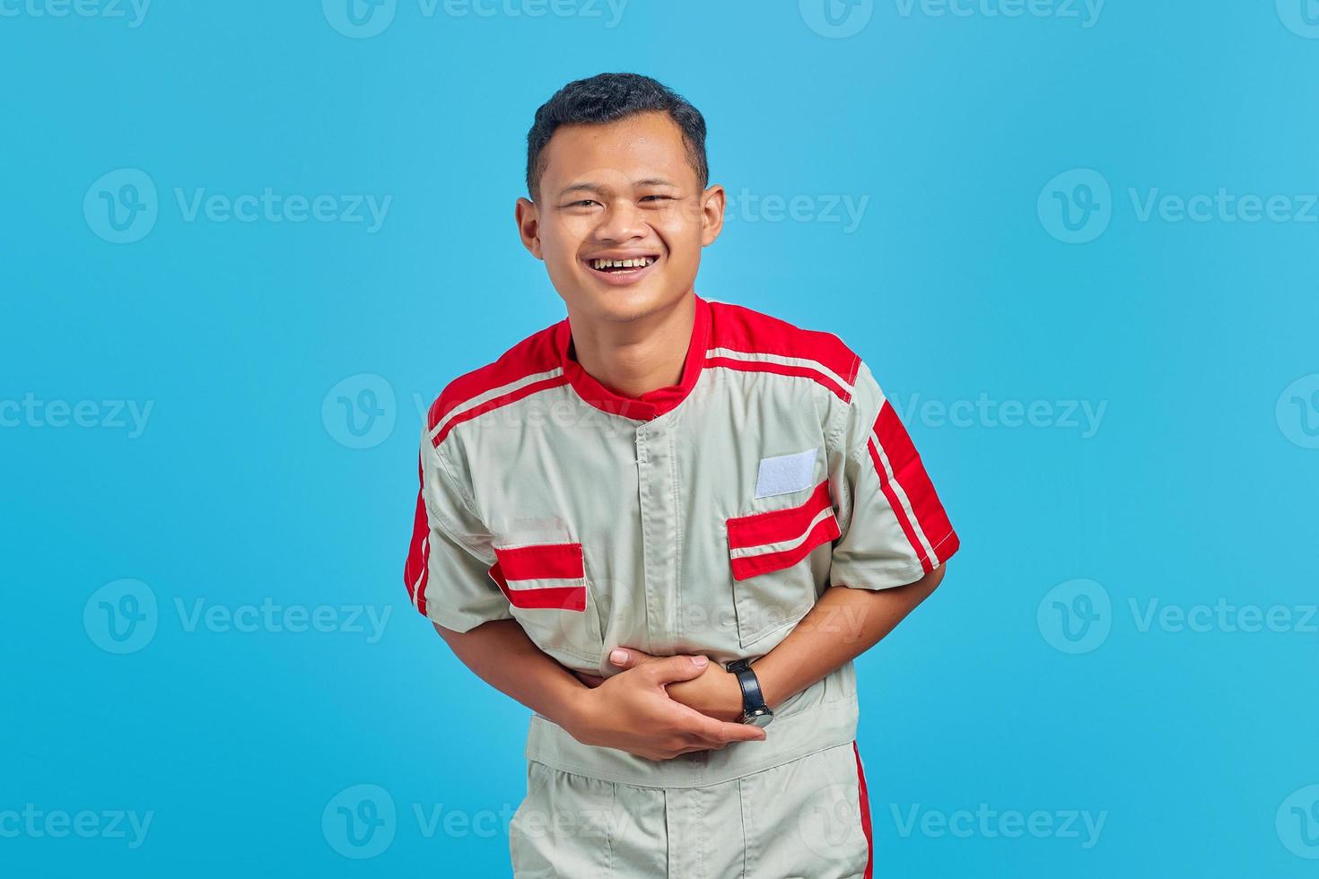 Portrait of young Asian mechanic showing loud laughter with hands on stomach over blue background photo