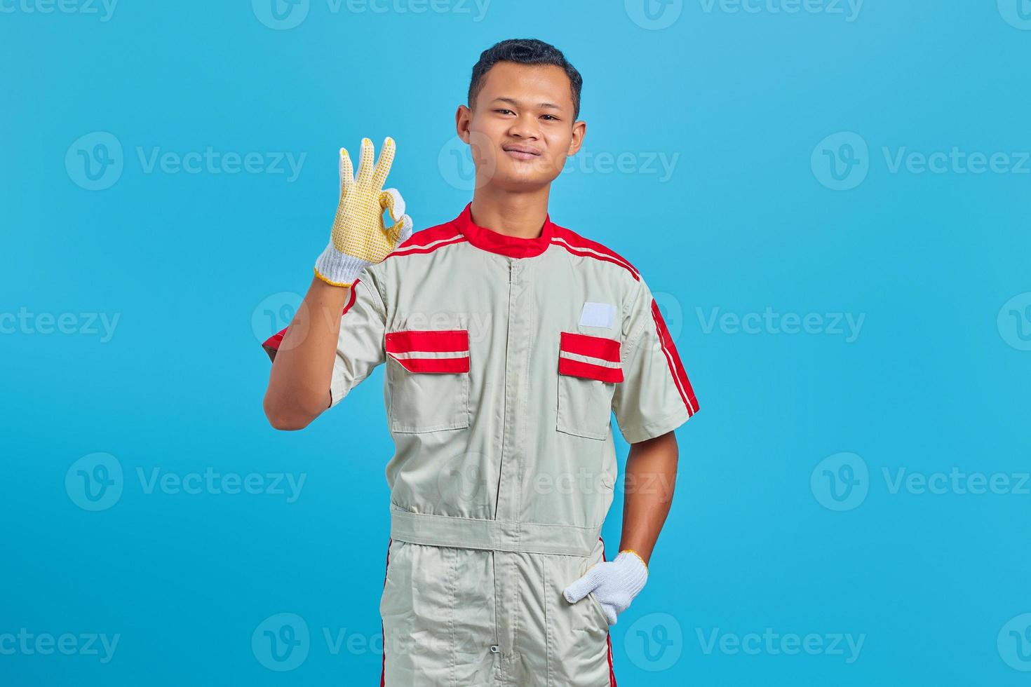 Portrait of smiling young Asian mechanic making okay gesture, showing approval symbol isolated on blue background photo