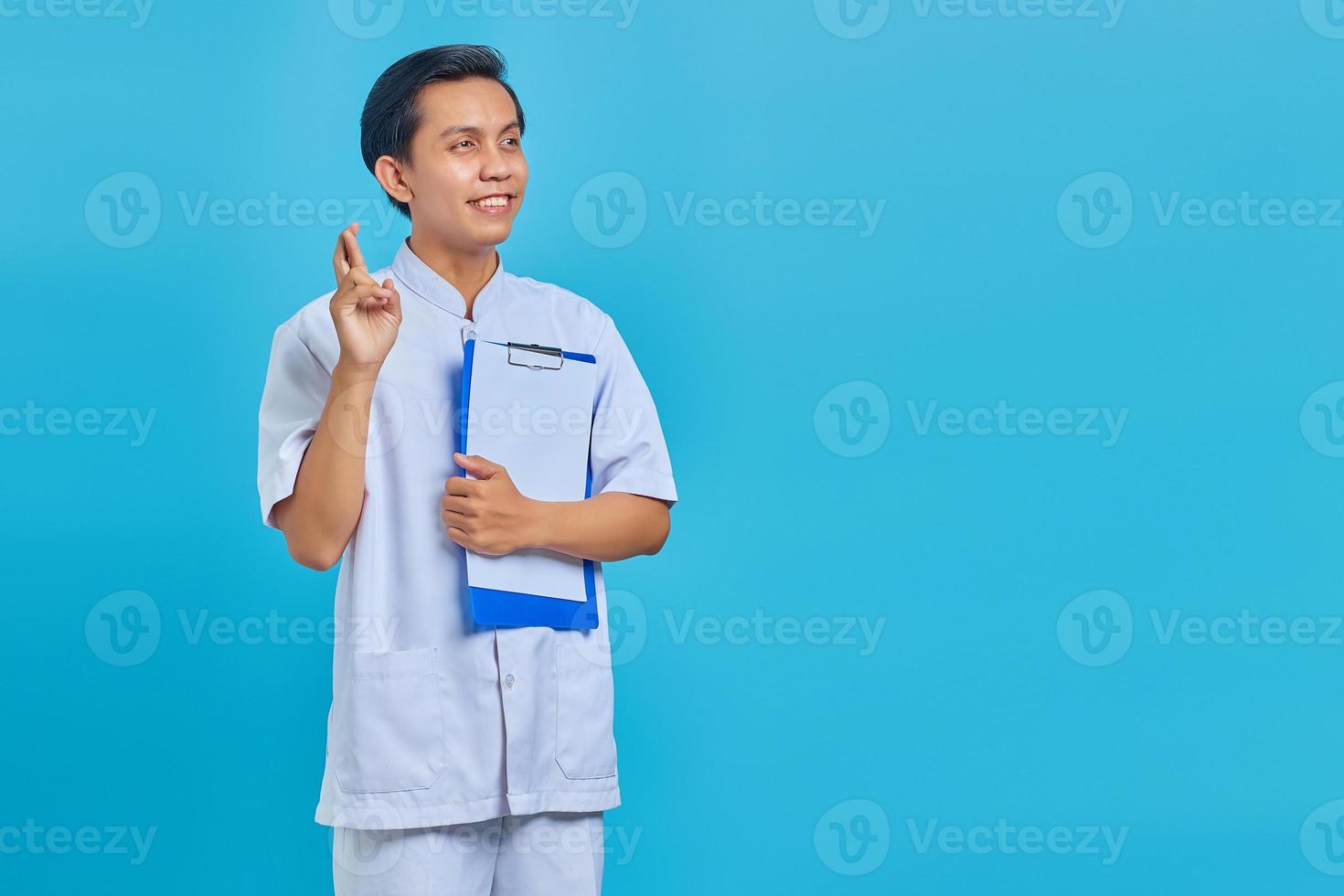 Portrait of happy young Asian male nurse crossing fingers and showing clipboard on blue background photo