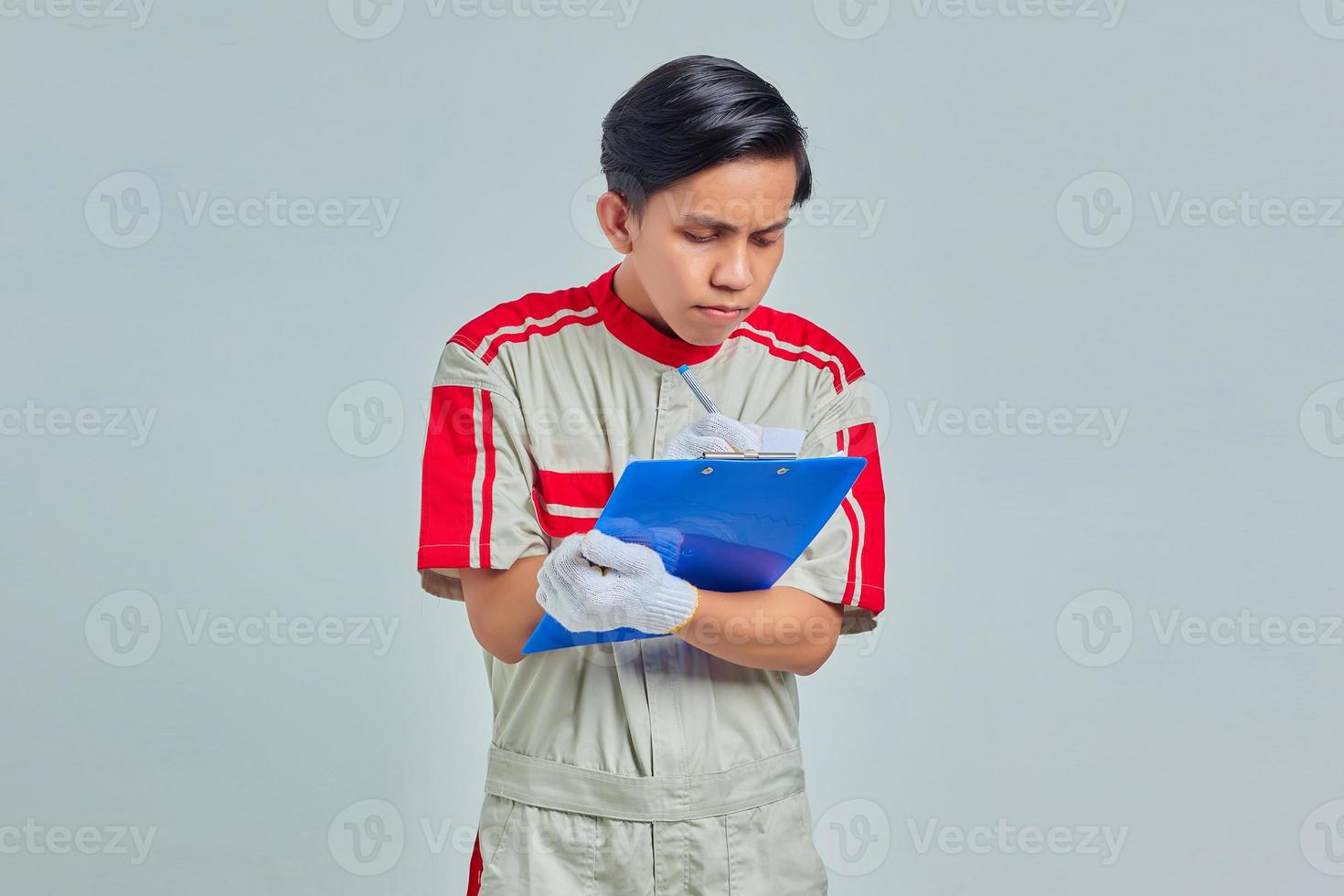 Sonriente joven mecánico tomando notas en el portapapeles sobre fondo gris foto