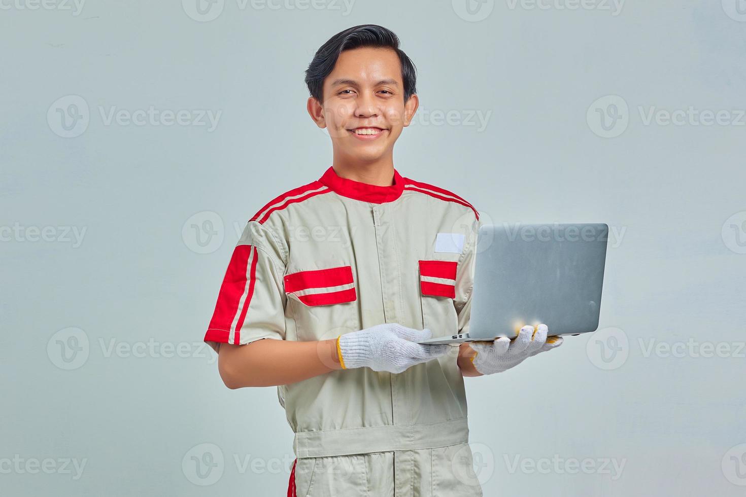 Sonriente joven guapo mecánico hombre vestido con uniforme sosteniendo portátil y mirando a cámara aislada sobre fondo gris foto