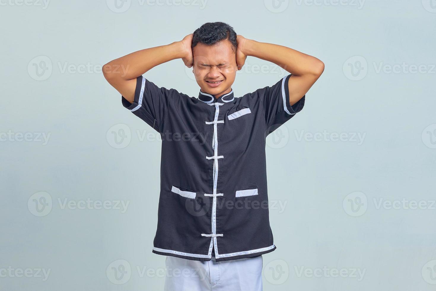 Portrait of frustrated Asian young man wearing taekwondo kimono covering ears with palms isolated on gray background photo