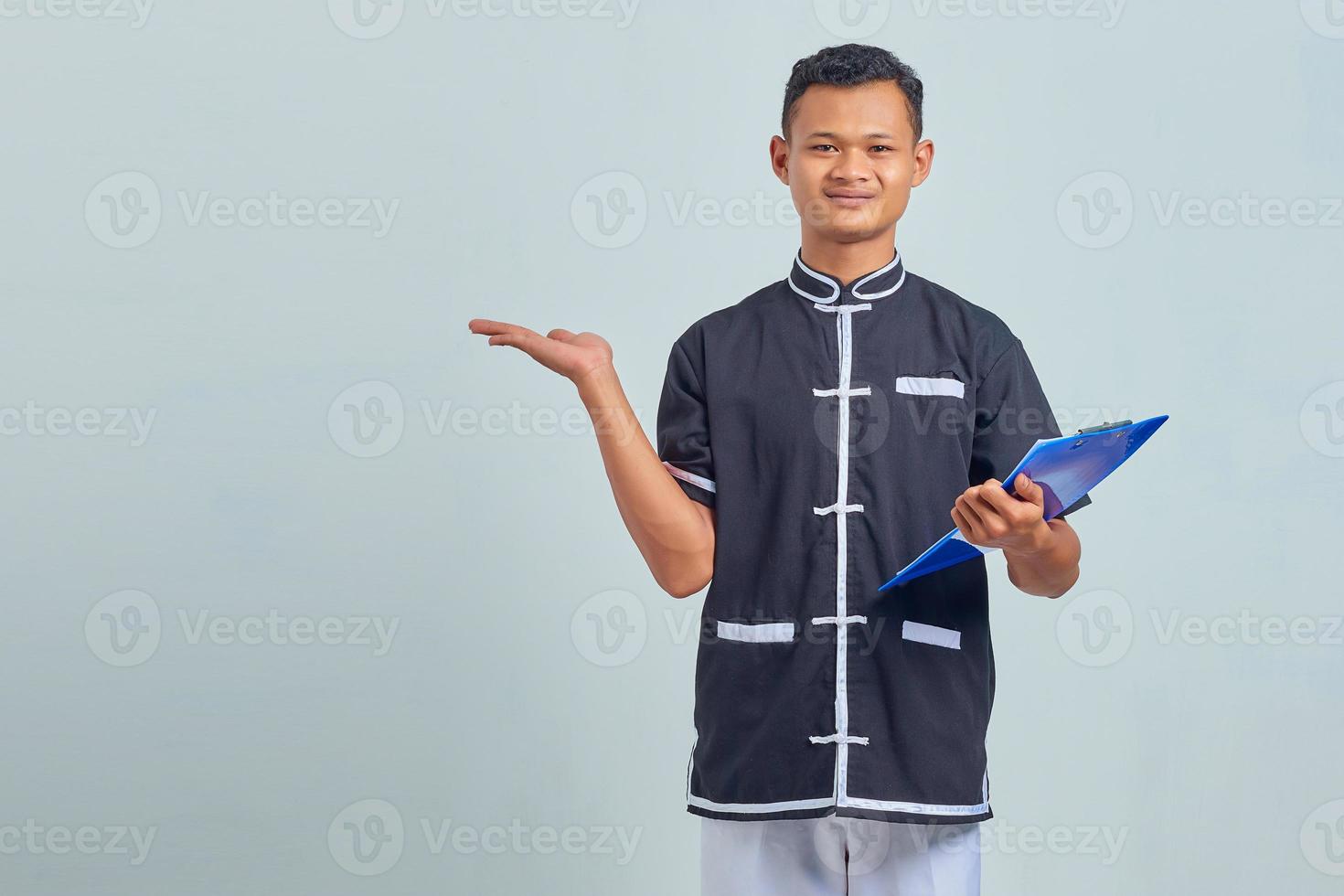 Portrait of smiling Asian young man wearing karate uniform holding clipboard and pointing sideways with palms photo