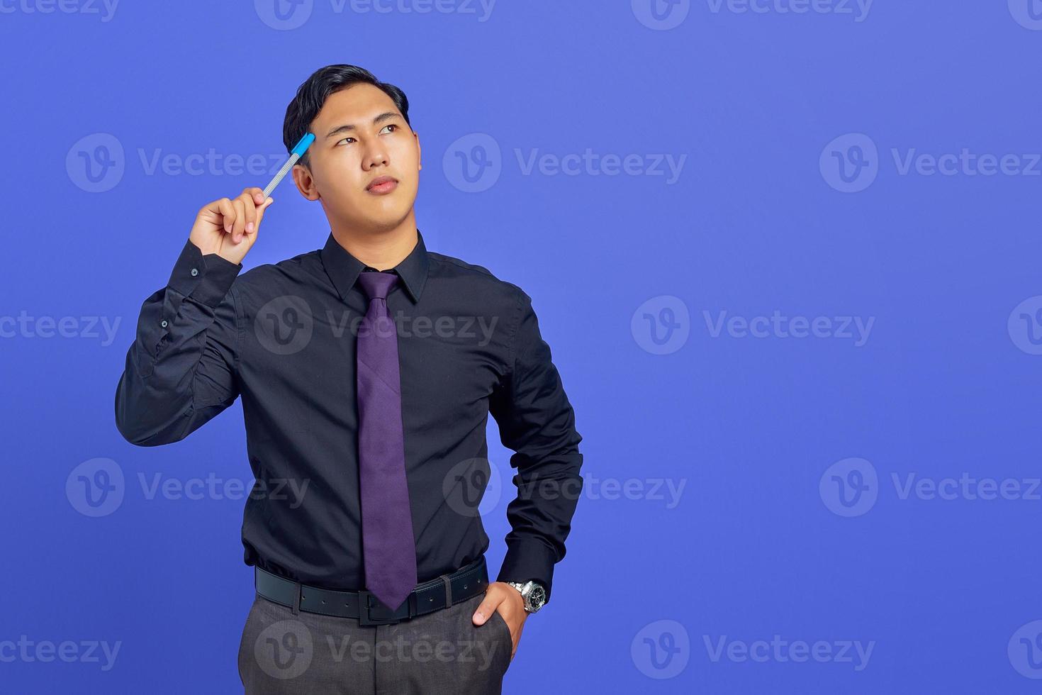 serio, joven, guapo, hombre de negocios, pensamiento, en, algo, y, tenencia, bolígrafo, sobre fondo púrpura foto