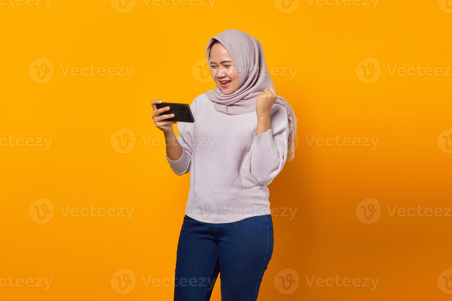 Portrait of happy young asian woman playing video game on mobile phone and hand gesture success on yellow background photo