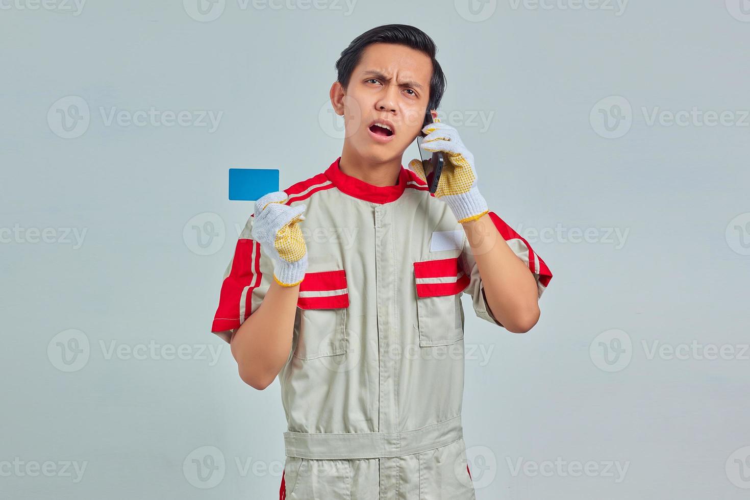 Shocked handsome mechanic talking on smartphone and holding credit card on gray background photo