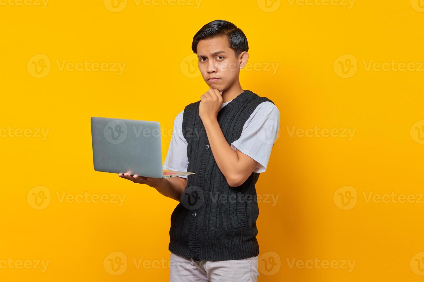 Portrait of confident Asian man holding laptop and looking at camera over yellow background photo
