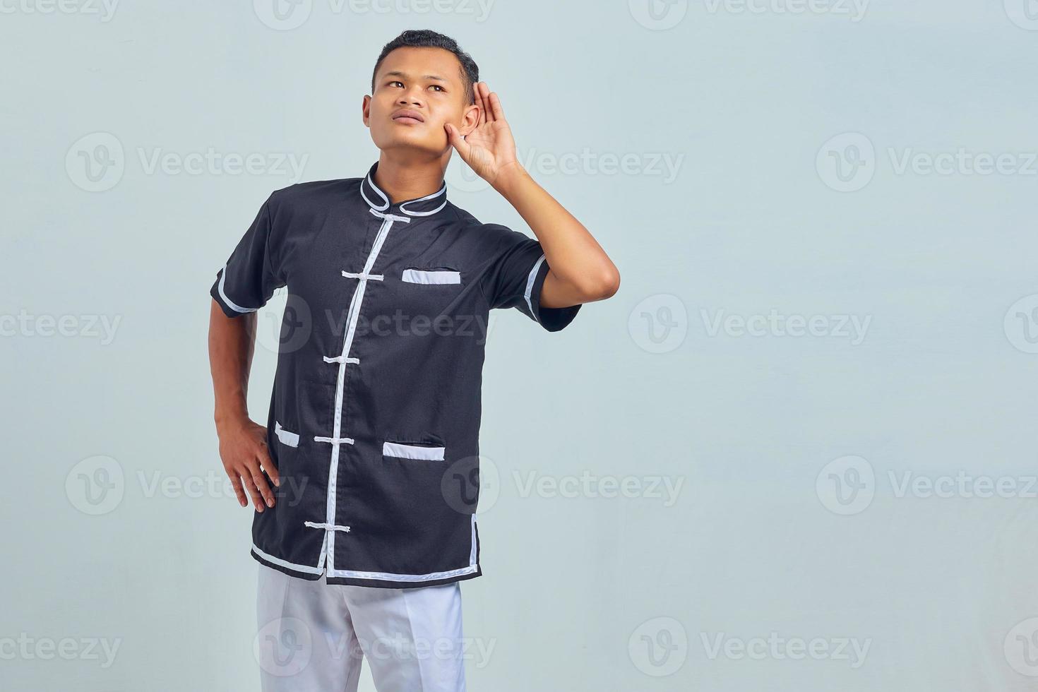 Portrait of cheerful asian young man wearing karate uniform trying to overhear secret conversation over gray background photo