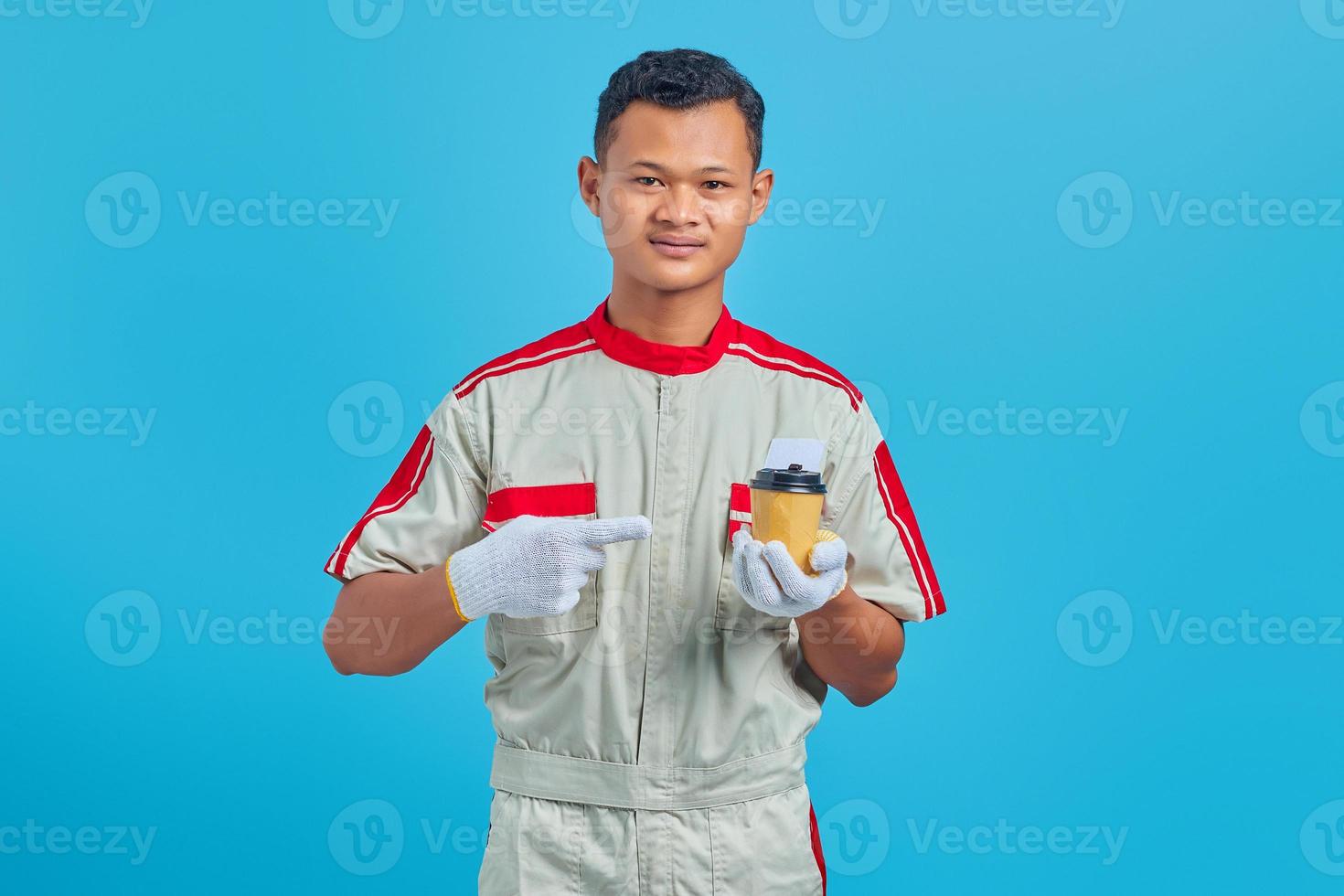 Retrato de sonriente joven mecánico asiático llevando una taza de café en la mano sobre fondo azul. foto