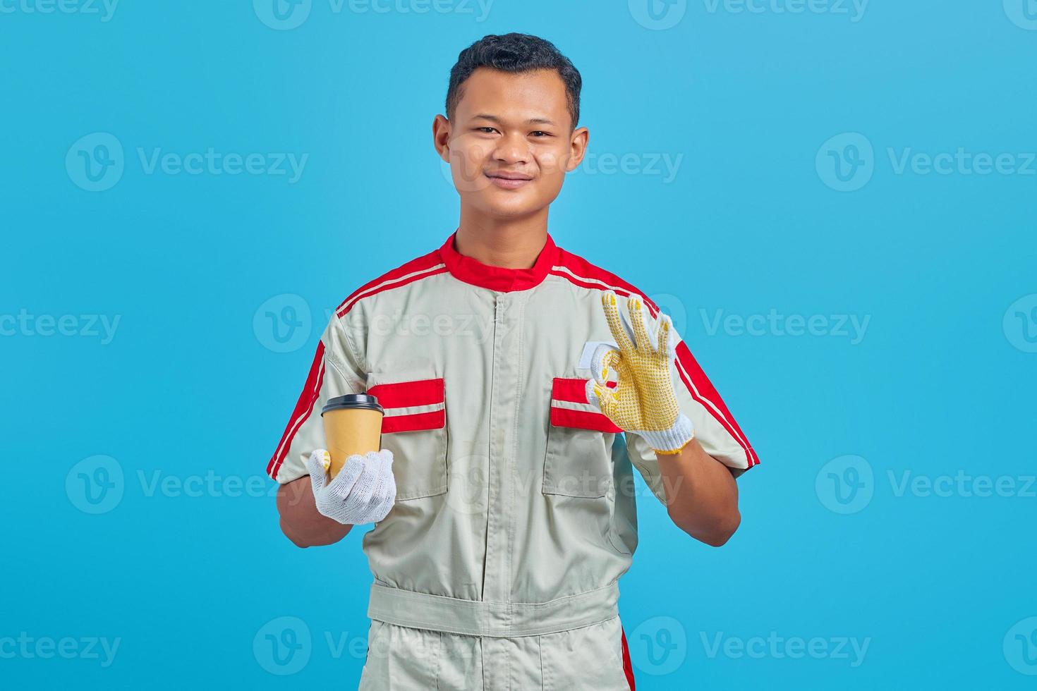 Retrato de sonriente joven mecánico asiático sosteniendo una taza de café en la mano y mostrando una buena actitud sobre fondo azul. foto